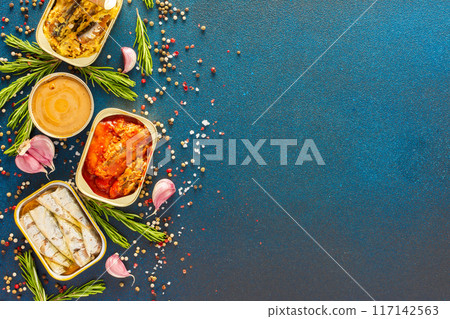 Different open tin cans with canned fish like mackerel, sprat, sardine, fish pate among spices and herbs on a blue background, viewed from the top 117142563