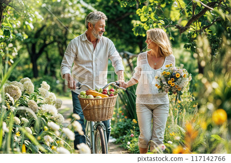 Senior couple walking in park with bike, picnic basket with harvest fruits and vegetables, flowers. Happy mature people with bicycle. Adult family outside. Woman with summer flower, man on nature park 117142766