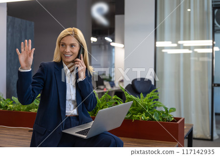 Businesswoman calling talking on mobile phone while working on laptop in office 117142935