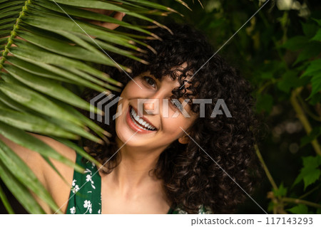Happy woman with curly hair among tropical leaves in wellness summer atmosphere 117143293