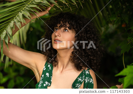 Winsome woman with curly hair and fresh greenery in the background Winsome woman with curly hair and fresh greenery in the background 117143303