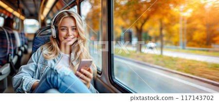 Woman in bus with phone. Girl in transport with smartphone and headphones. Young people inside train or tram. Happy person with music travel to city. Student passenger with mobile. Autumn window view 117143702