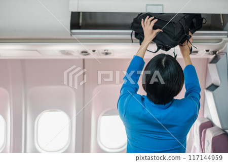 Flight attendant in blue uniform placing a black bag in the overhead compartment of an airplane cabin. 117144959