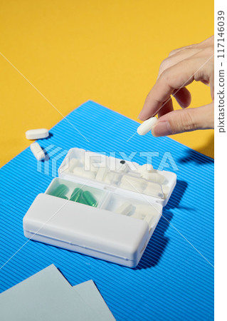 High angle close up shot at the colorful flat with a white daily pill box displayed in the center, a beautiful hand hold a white tablet in space. Blank space for product presentation, copy space 117146039