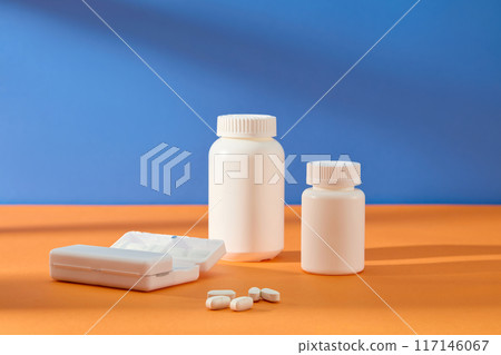 White unlabeled medicine jars and white pill box displayed in the center and standing out on blue and orange background. Frontal shot, blank space for advertising White unlabeled medicine jars and white pill box displayed in the center and standing out on blue and orange background. Frontal shot, blank space for advertising 117146067