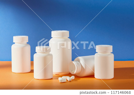 Different sizes unbranded medicine bottles in white displayed on orange countertop with some white tablets fall out in front, over blue background. Front angle shot, empty space for text Different sizes unbranded medicine bottles in white displayed on orange countertop with some white tablets fall out in front, over blue background. Front angle shot, empty space for text 117146068
