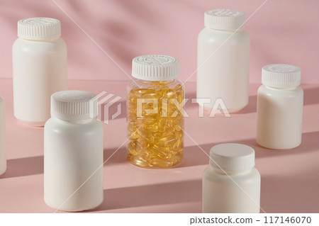 Neatly arranged medicine bottles mockup on pink counter top, transparent supplement bottle filled with yellow vitamin capsule surrounded in center by white unbranded bottles. High angle, copy space Neatly arranged medicine bottles mockup on pink counter top, transparent supplement bottle filled with yellow vitamin capsule surrounded in center by white unbranded bottles. High angle, copy space 117146070