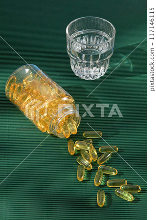 The template to design promotional pic with a transparent bottle filled by yellow supplement capsules flat lay on green countertop, some capsule poured from which. A glass of water placed by side The template to design promotional pic with a transparent bottle filled by yellow supplement capsules flat lay on green countertop, some capsule poured from which. A glass of water placed by side 117146115