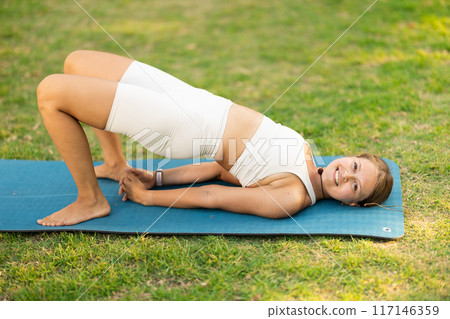 Girl in park on green grass of lawn performs Setu Bandha Sarvangasana 117146359