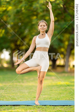 Girl doing Vrikshasana balancing yoga asana on green meadow Girl doing Vrikshasana balancing yoga asana on green meadow 117146402