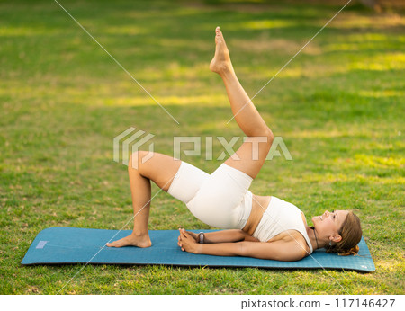 Girl in park on green grass of lawn performs Setu Bandha Sarvangasana 117146427