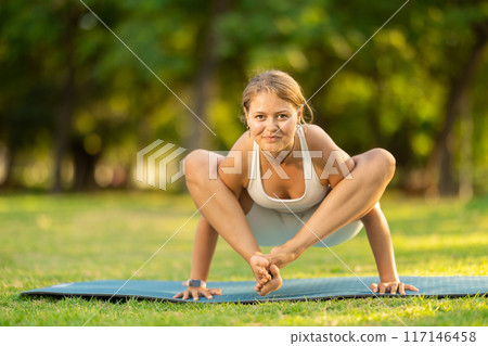 Girl performing shoulder-pressing pose Bhujapidasana in summer park 117146458