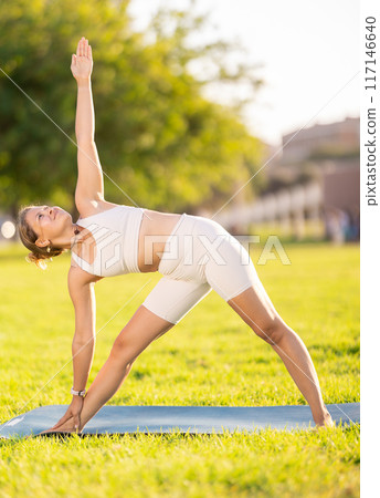 Girl in park on green grass of lawn performs Trikonasana, triangle asana Girl in park on green grass of lawn performs Trikonasana, triangle asana 117146640