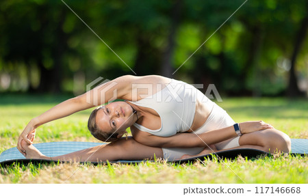Girl in park on green grass of lawn performs Parivritta Janu Shirshasana 117146668