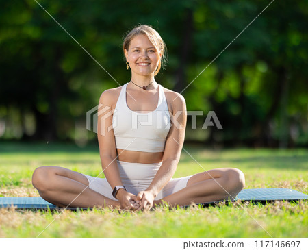 Smiling girl performing butterfly pose on exercise mat in summer park 117146697