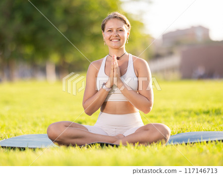 Smiling girl meditating in lotus position on green sunlit meadow 117146713