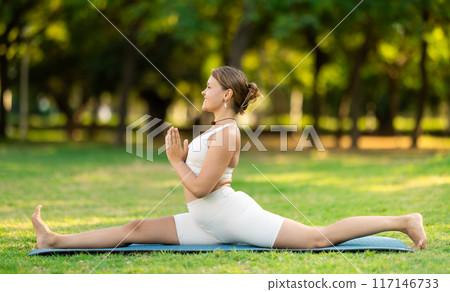 Fit girl performing front split with hands in namaste in summer park Fit girl performing front split with hands in namaste in summer park 117146733