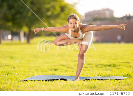 Girl in park on green grass of lawn performs Garudasana 117146763