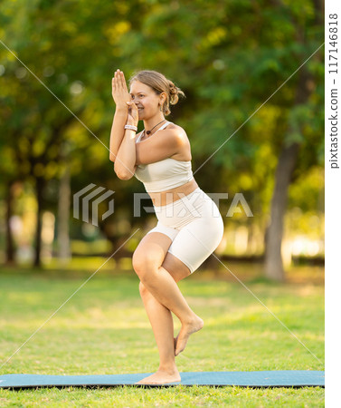 Girl in park on green grass of lawn performs Garudasana 117146818