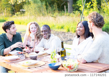 A group of friends is happily enjoying a meal together outdoors in the sunshine on a summer day 117148201
