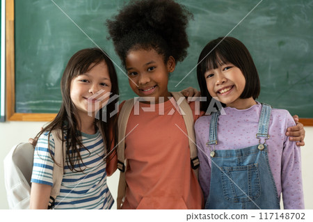 Three diverse and happy school girls standing together in a classroom with a chalkboard behind them, smiling and showing their friendship and unity with backpacks Three diverse and happy school girls standing together in a classroom with a chalkboard behind them, smiling and showing their friendship and unity with backpacks 117148702