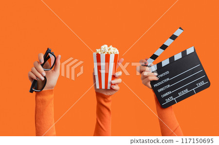 Three womans hands holding clapperboard, popcorn and 3d glasses on an orange background 117150695