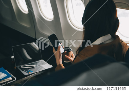 A young Asian woman, seated as an airplane passenger, contemplates dollar bills, reflecting on lucrative opportunities and aspirations towards billionaire status. 117151665