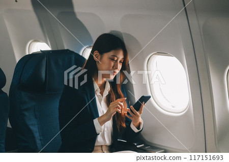 A young Asian female airplane passenger sits by the window during the flight, holding her boarding pass, with her carry-on luggage stored overhead, wearing her seatbelt, and enjoying the journey. A young Asian female airplane passenger sits by the window during the flight, holding her boarding pass, with her carry-on luggage stored overhead, wearing her seatbelt, and enjoying the journey. 117151693