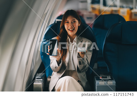 A young Asian woman, seated as an airplane passenger, contemplates dollar bills, reflecting on lucrative opportunities and aspirations towards billionaire status. 117151743