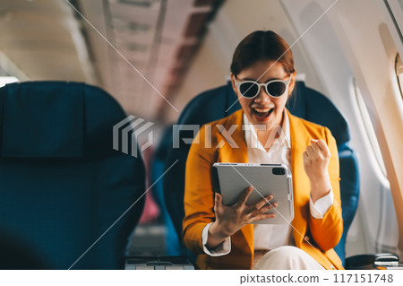A young Asian woman, seated as an airplane passenger, contemplates dollar bills, reflecting on lucrative opportunities and aspirations towards billionaire status. 117151748