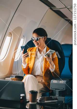 A young Asian woman, seated as an airplane passenger, contemplates dollar bills, reflecting on lucrative opportunities and aspirations towards billionaire status. A young Asian woman, seated as an airplane passenger, contemplates dollar bills, reflecting on lucrative opportunities and aspirations towards billionaire status. 117151750