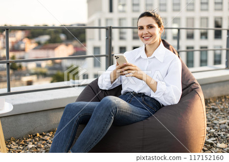 Smiling woman using smartphone on rooftop outdoor chair 117152160