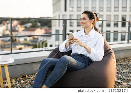 Woman relaxing on rooftop with smartphone in hand smiling 117152174