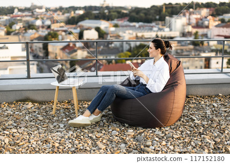 Woman working remotely from rooftop with laptop and phone 117152180