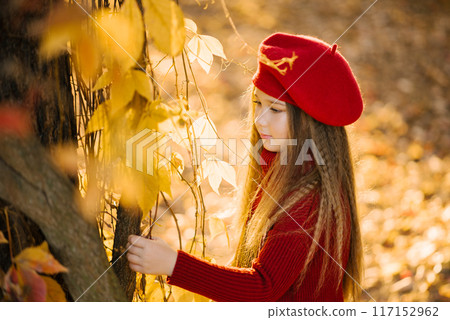 Cute girl in a burgundy beret stands near a tree in autumn b touches a yellow leaf 117152962