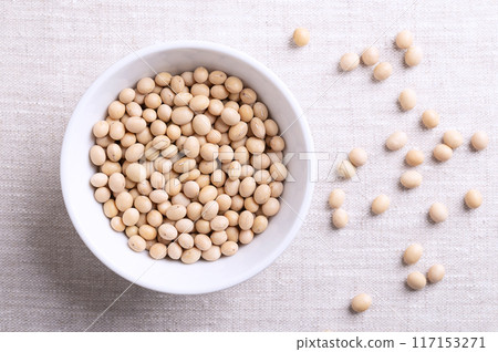 Dried soybeans, also soy beans or soya beans in a white bowl on linen fabric. Whole and raw seeds of the legume and oilseed Glycine max, with a very high protein and oil content. Close-up from above. 117153271