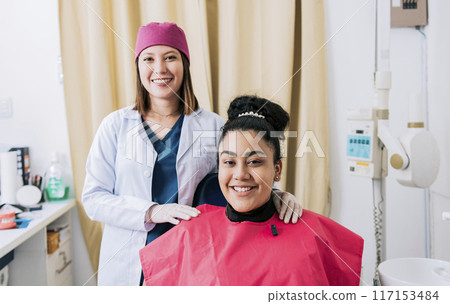 Female dentist with satisfied patient smiling at camera, Female dentist with patient smiling friendly. Portrait of dentist with patient smiling at camera in office 117153484