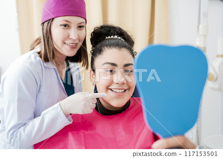 Dentist with satisfied patient smiling at dental mirror, Satisfied patient in dental clinic looking at mirror, female patient checking teeth after curing teeth in dental clinic. Dentist with satisfied patient smiling at dental mirror, Satisfied patient in dental clinic looking at mirror, female patient checking teeth after curing teeth in dental clinic. 117153501
