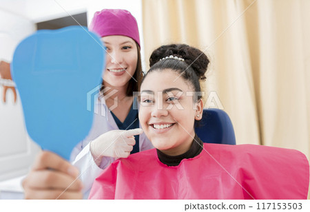 Satisfied patient in dental clinic looking at mirror, dentist next to satisfied patient smiling at dental mirror, female patient checking teeth after curing teeth in dental clinic. Satisfied patient in dental clinic looking at mirror, dentist next to satisfied patient smiling at dental mirror, female patient checking teeth after curing teeth in dental clinic. 117153503