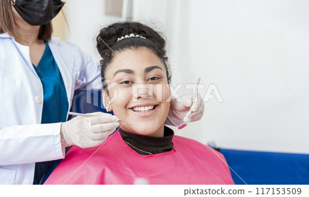 Dentist doctor examining mouth to female patient looking at camera lying on chair, Female dentist with probe and dental mirror examining mouth to lying patient. Health and dental care concept 117153509