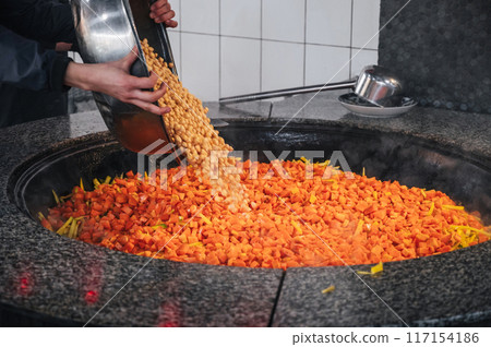 male chef adds chickpeas to a cauldron with carrots to cook traditional oriental Uzbek pilaf in asian kitchen at a restaurant in Uzbekistan male chef adds chickpeas to a cauldron with carrots to cook traditional oriental Uzbek pilaf in asian kitchen at a restaurant in Uzbekistan 117154186