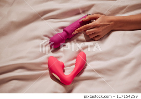 sex vibrator and vacuum stimulator in a woman girl hand on a white sheet on bed in bedroom. Sex toys for female satisfaction and orgasm sex vibrator and vacuum stimulator in a woman girl hand on a white sheet on bed in bedroom. Sex toys for female satisfaction and orgasm 117154259