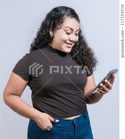 Smiling latin girl using cell phone isolated. Cheerful young woman with curly hair texting on cell phone isolated. 117154519