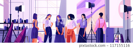 Fashion photoshoot scene with models and photographers in a studio setting with lighting equipment shelves and a large window 117155116