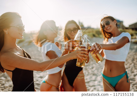 Nice girls cheers and drink beers on the beach at sunset. Young four girl in bikini enjoying on beach holiday. 117155138