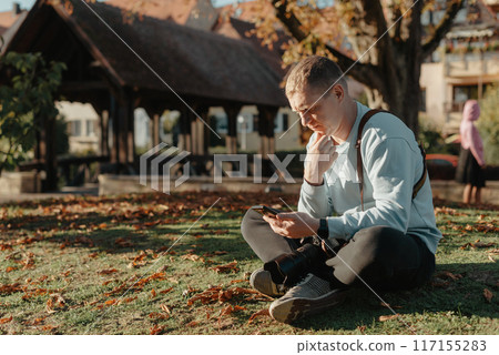 Professional Photographer Taking Picture Of Beautiful Autumn Park. Man Professional Photographer Sit With Camera And With Smartphone In Autumn Park. Retouched, Vibrant Colors, Brownish Tones. 117155283