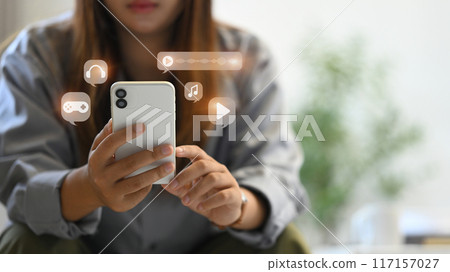 Young woman using smartphone, interacting with various multimedia icons including music, video and gaming 117157027