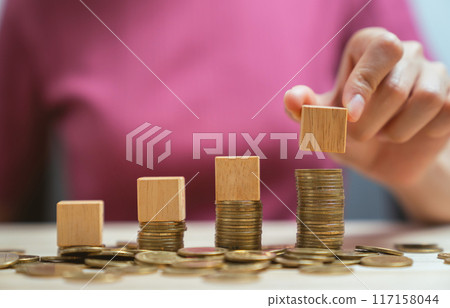 Hand holding wooden cubes on a pile of gold coins. 117158044