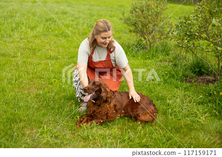 Cheerful happy young woman playing with her dog in the yard of the house in summer. Beautiful Irish Setter dog is lying in grass. 30s adult girl adopting adorable dog in animal shelter Cheerful happy young woman playing with her dog in the yard of the house in summer. Beautiful Irish Setter dog is lying in grass. 30s adult girl adopting adorable dog in animal shelter 117159171