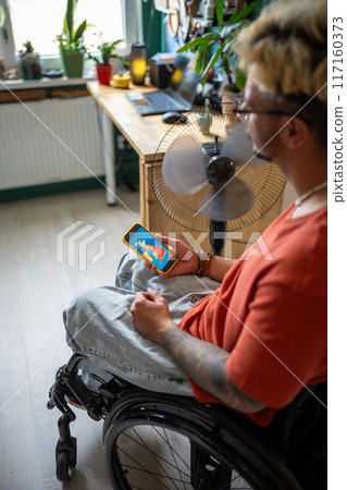 Disabled man in wheelchair looks at weather forecast on phone screen, sits by fan at home. Heat wave 117160373